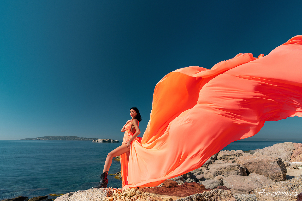 A woman in a flowing orange dress stands confidently on rocky cliffs with the deep blue sea and distant islands of Kavouri, Athens in the background, creating a vivid and striking scene.